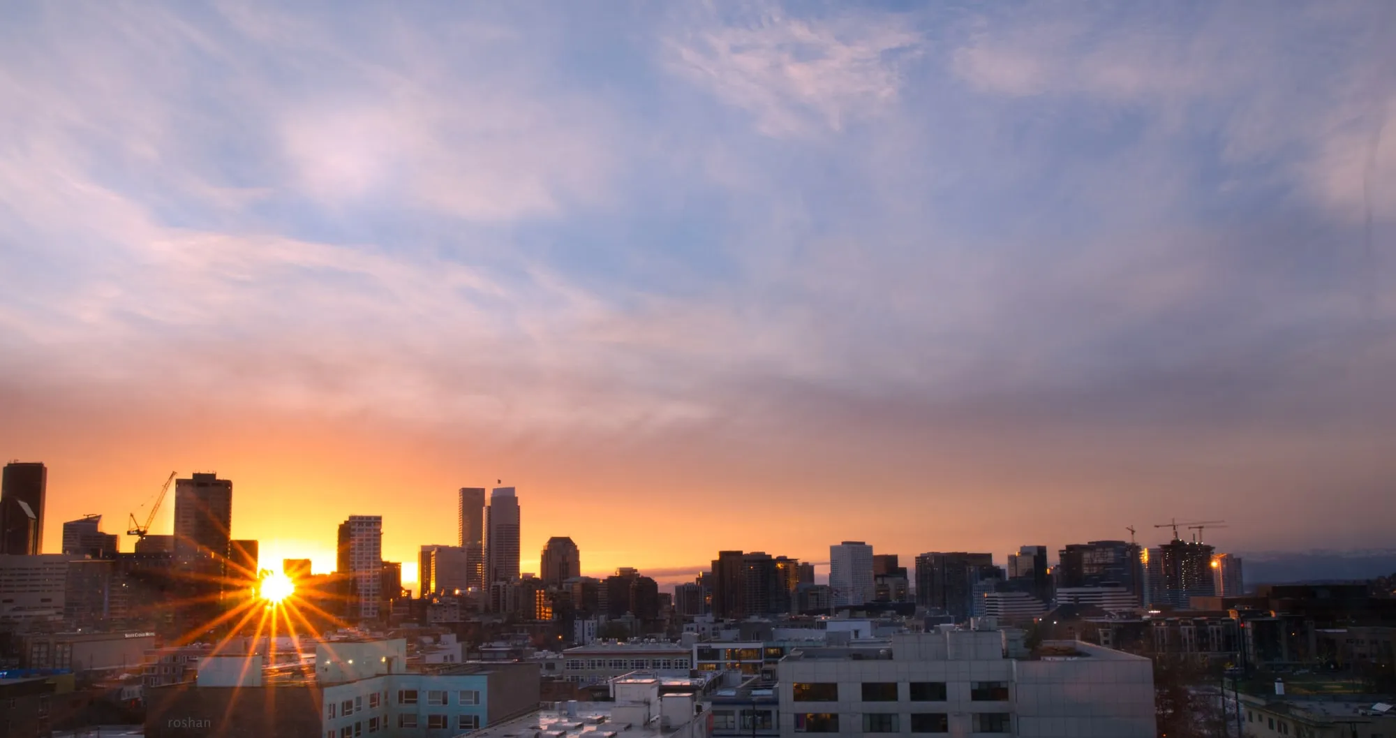 A vivid Seattle sunset with sun rays breaking between downtown buildings, mountains in the distance.